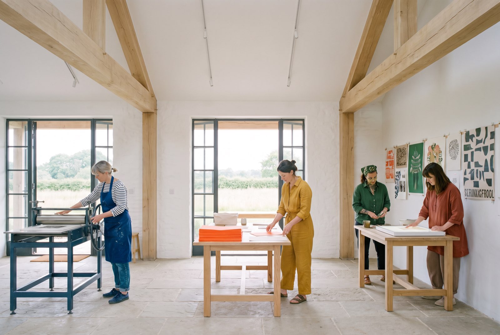 Four people work side by side in a timber-beamed studio: one at a print press, the others at oak tables with paper and prints. Bold prints are pinned to the white wall and a stack of orange paper sits on the middle table.