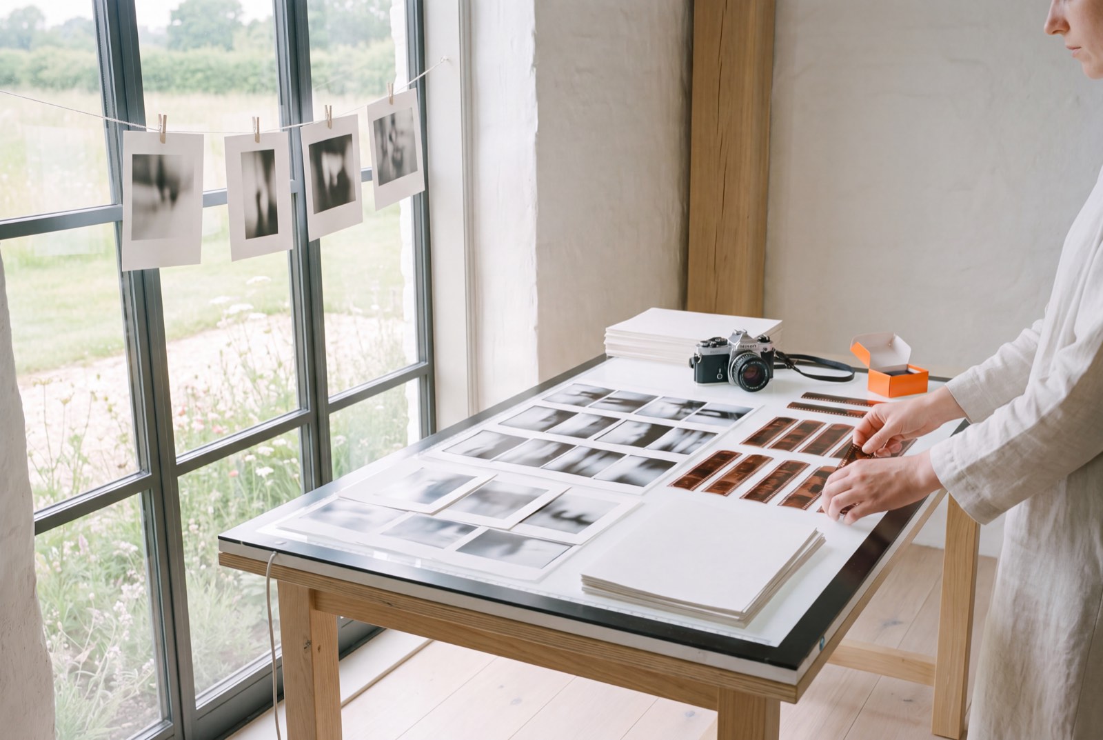 Contact sheets laid out on a light table, with black-and-white prints pinned above and a film camera nearby.