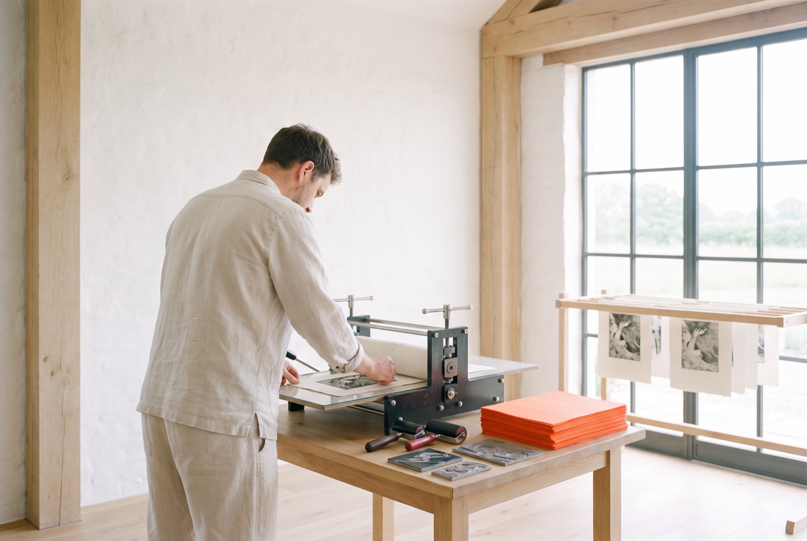 A printmaker pulls a print on a small press, with fresh prints drying on a rack above.