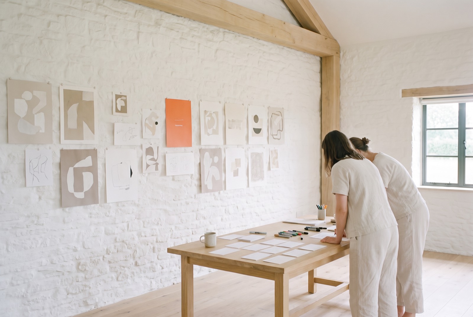 Two people study layouts at a table beside a wall covered in pinned sketches and ideas.