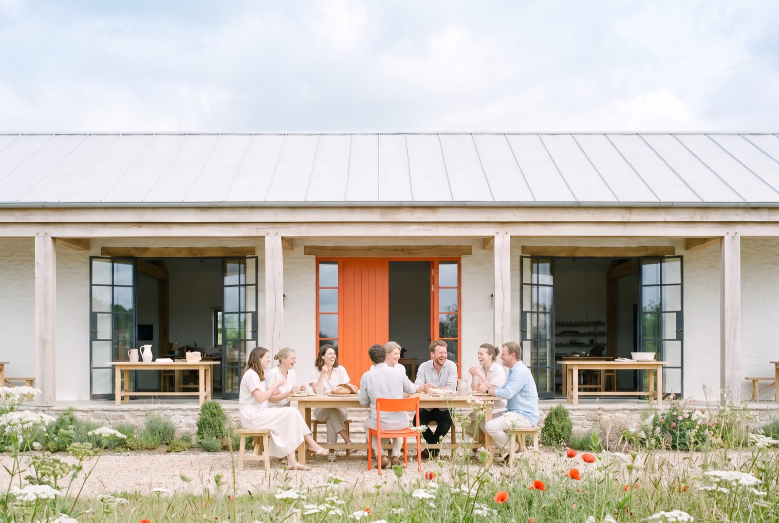 Eight people share a long lunch at a wooden table in front of the barn, talking and laughing. An orange door and an orange chair stand out, with wildflowers and poppies in the foreground.