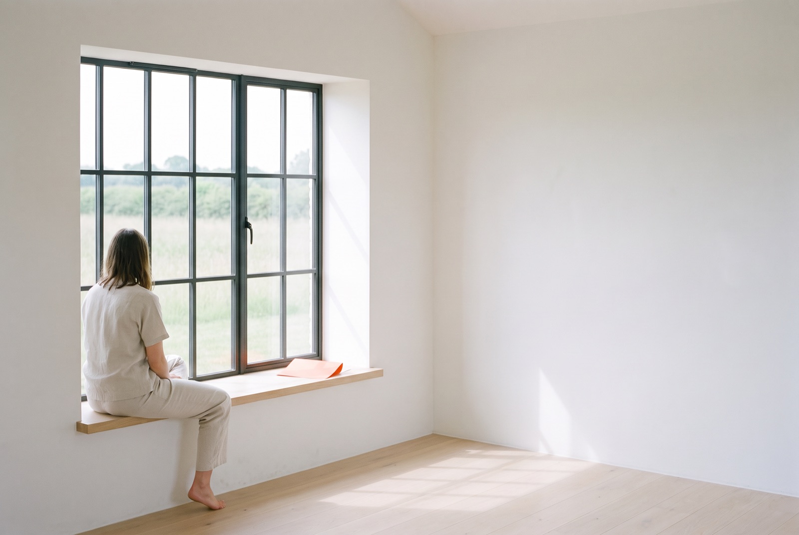 One person sits on a wide oak window seat, looking out at the meadow through a steel-framed window. An orange sheet of paper lies beside them in a quiet white room.