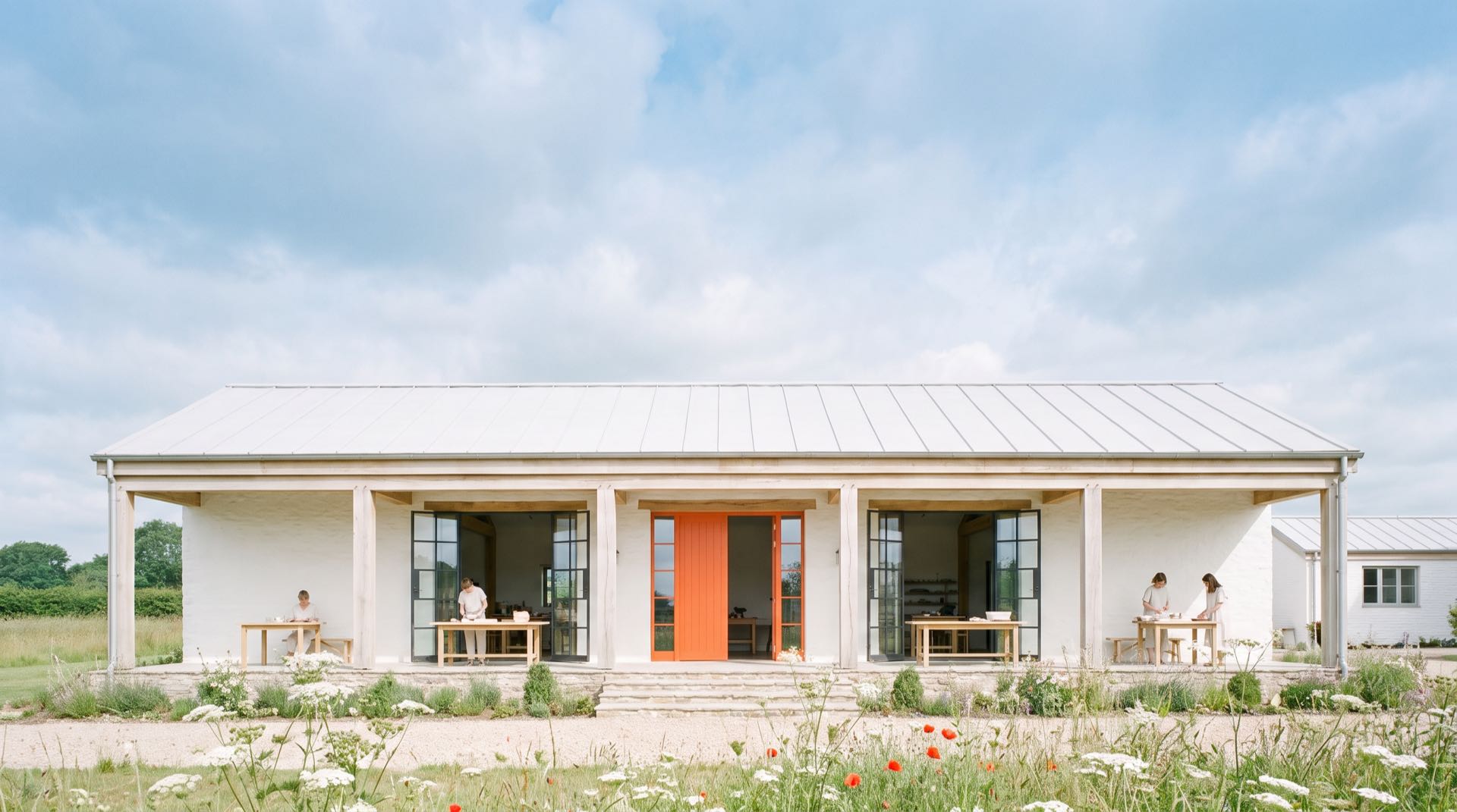 The whitewashed barn seen from the meadow under a wide sky, with an orange front door, open studios on either side and a few people working at tables under the porch.